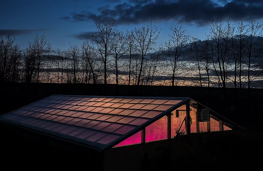 Vue extérieure d’une serre Walipini semi‑enterré en Alberta (Canada), photographié à l’automne. Le toit en polycarbonate triple paroi laisse filtrer la lumière des lampes horticoles. Cette ambiance met en valeur l’atmosphère chaleureuse et humide du refuge, alors que les températures extérieures peuvent descendre jusqu’à –40 °C.